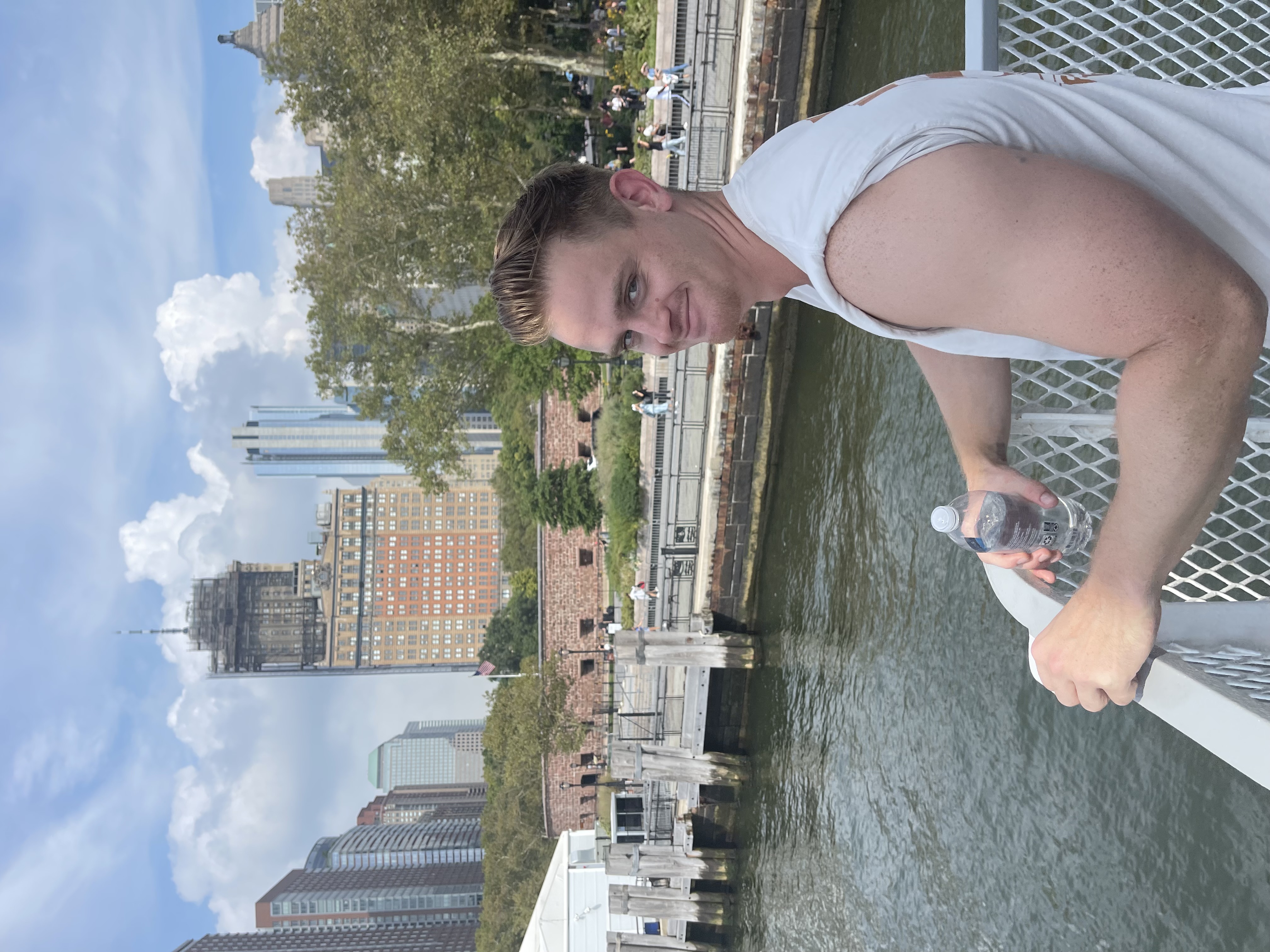 Smiling person leaning on a ferry railing with a city skyline in the background, holding a water bottle.