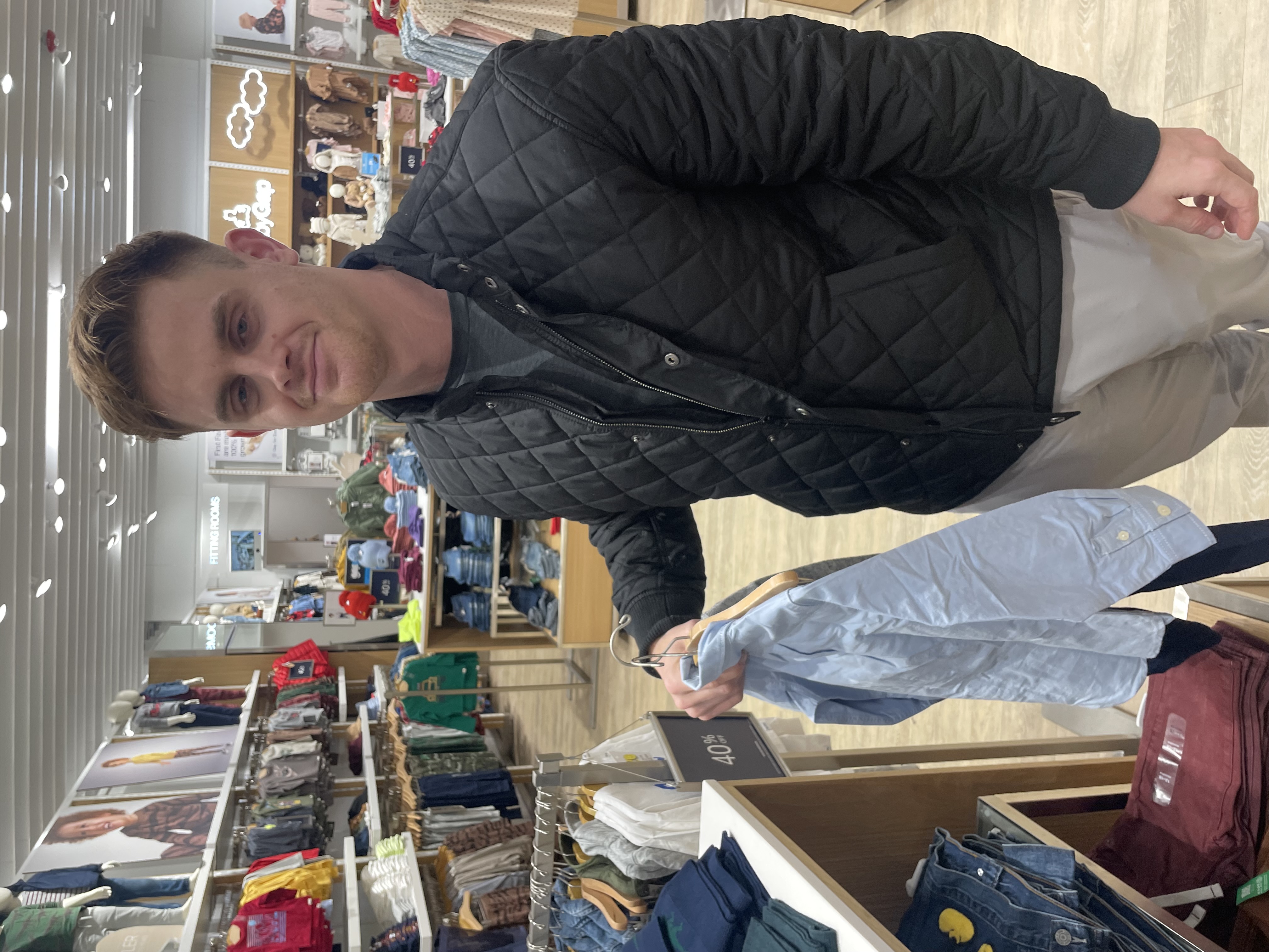 Person in a quilted jacket holding up a tiny button-down shirt inside a clothing store.