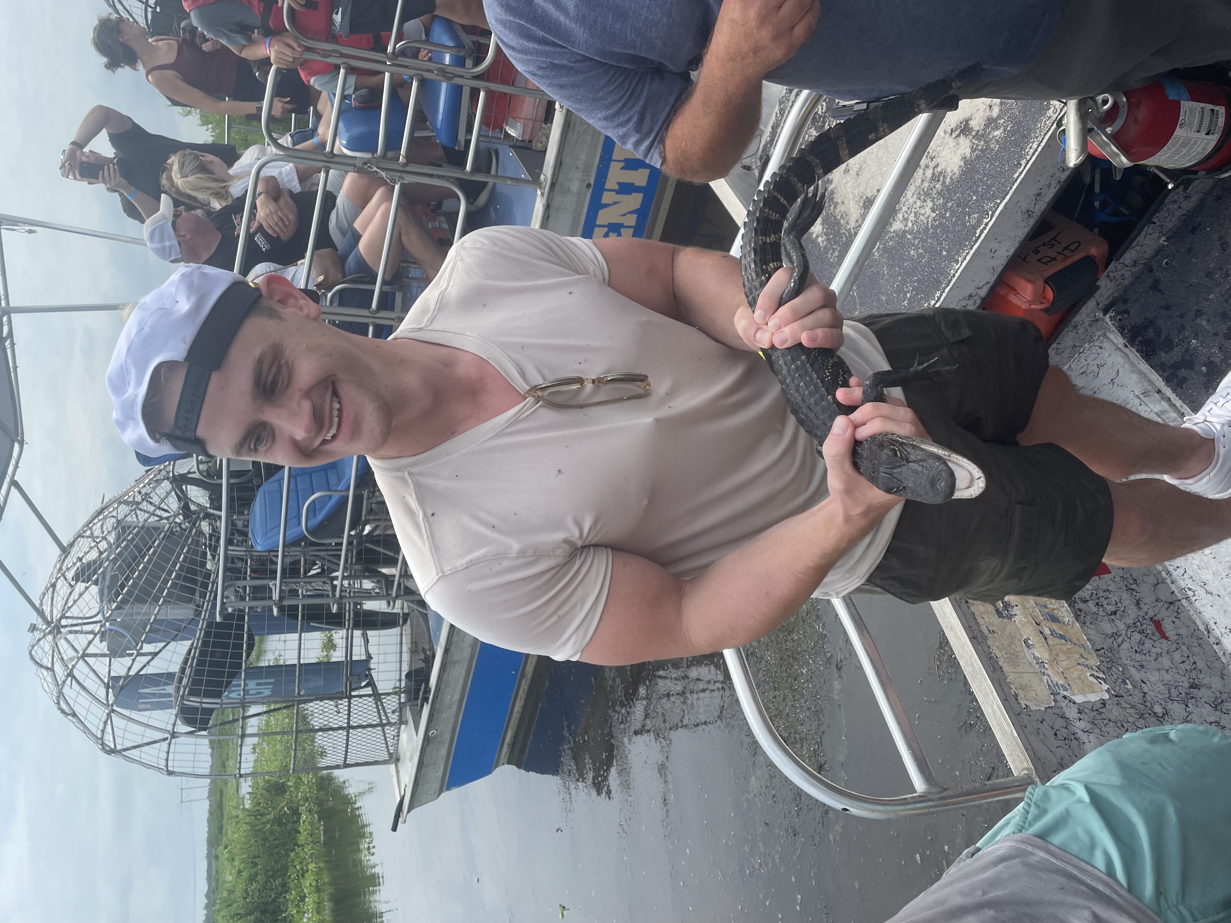Person holding a small alligator on an airboat tour while smiling at someone nearby.